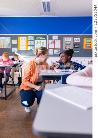 In school, female teacher kneeling and helping african american girl with classwork at desk 125353044