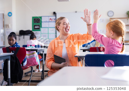 In school, female teacher high-fiving girl in classroom, holding tablet, smiling 125353048