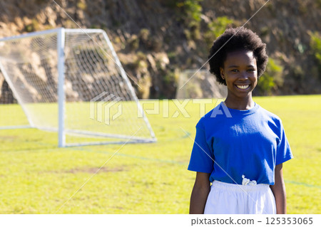 Smiling african american girl standing on soccer field with goal post, copy space, at school 125353065