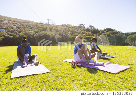 In school, diverse girls sitting on yoga mats stretching legs outdoors on grass In school, diverse girls sitting on yoga mats stretching legs outdoors on grass 125353082