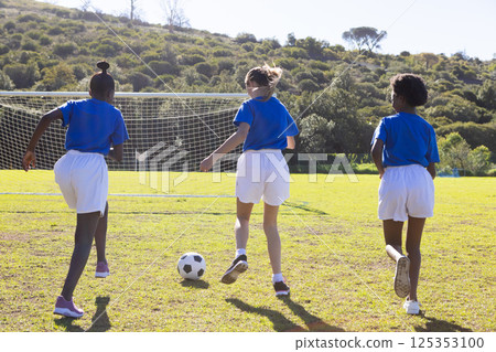 Playing soccer, three school diverse girls running towards goal on sunny field 125353100
