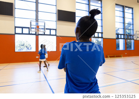 Watching basketball game, african american girl in school gym observing players on court Watching basketball game, african american girl in school gym observing players on court 125353125