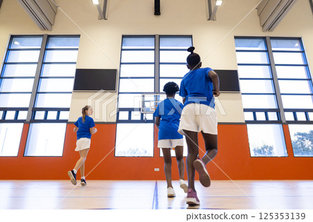 Playing basketball in school gym, diverse girls running towards hoop in uniforms Playing basketball in school gym, diverse girls running towards hoop in uniforms 125353139