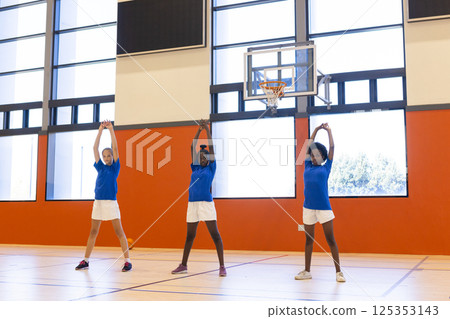 In school gym, diverse girls wearing uniforms stretching arms during physical education class In school gym, diverse girls wearing uniforms stretching arms during physical education class 125353143