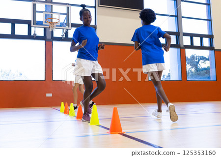 Running in school gym, diverse girls exercising around cones during physical education class 125353160