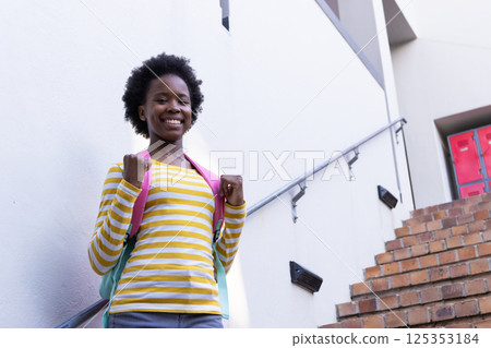 Smiling african american girl with backpack standing on school staircase, ready for class 125353184