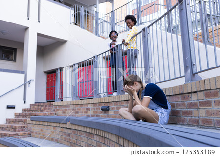 Crying girl sitting alone while two african american kids laughing at her in school Crying girl sitting alone while two african american kids laughing at her in school 125353189