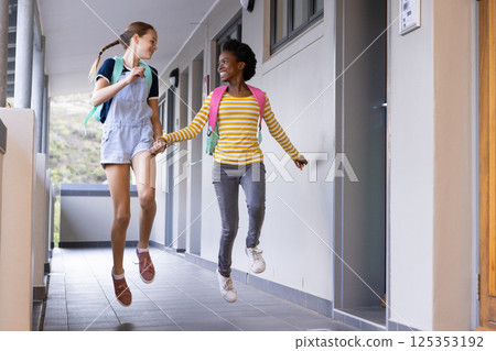 Jumping and holding hands, two diverse schoolgirls with backpacks walking in hallway Jumping and holding hands, two diverse schoolgirls with backpacks walking in hallway 125353192