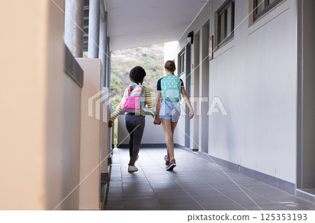 Walking in school hallway, two diverse girls with backpacks holding hands, heading to class 125353193