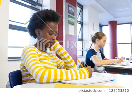 In school, african american girl writing in notebook while another student using laptop nearby 125353210