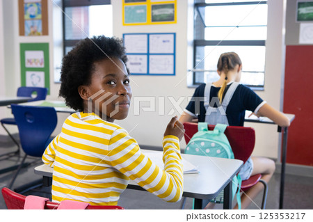 In school, african american girl in striped shirt sitting at desk, looking back and smiling In school, african american girl in striped shirt sitting at desk, looking back and smiling 125353217