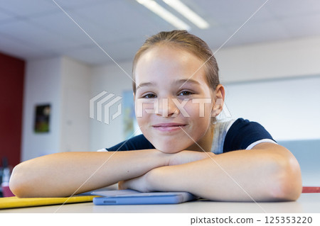 Smiling girl in school resting on desk with tablet, looking at camera 125353220