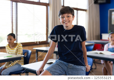 Smiling boy sitting on desk in classroom with students in background at school Smiling boy sitting on desk in classroom with students in background at school 125353225