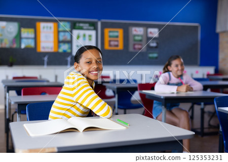 In school, girl smiling and sitting at desk with open book in classroom In school, girl smiling and sitting at desk with open book in classroom 125353231