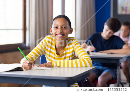 In school, girl smiling and writing in notebook at classroom desk In school, girl smiling and writing in notebook at classroom desk 125353232
