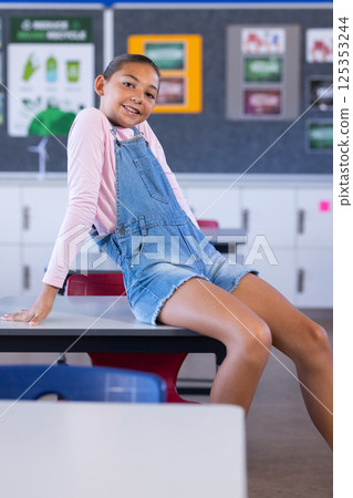 Smiling girl in overalls sitting on desk in school classroom, looking relaxed 125353244