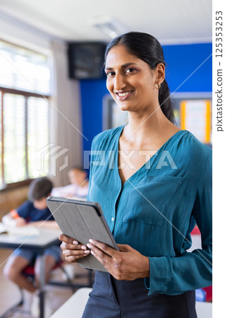 In school, Indian female teacher holding tablet and smiling in classroom with students 125353253