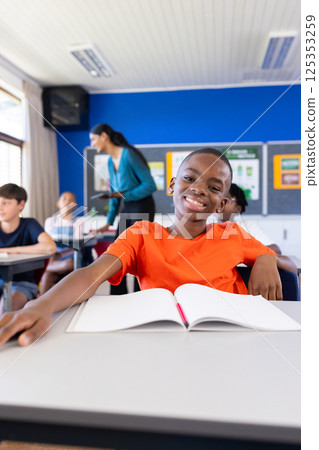Smiling boy in classroom with open notebook, surrounded by classmates in school 125353259
