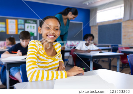 In school, smiling girl sitting at desk with classmates and female teacher in classroom, copy space 125353263