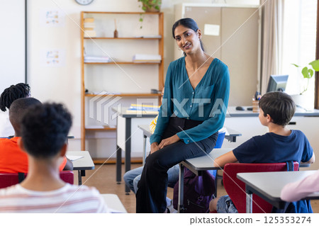 In school, Indian female teacher sitting on desk and engaging with students in classroom 125353274