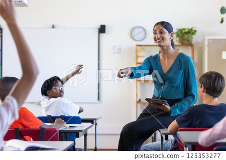 In school, Indian female teacher holding tablet and engaging with students raising hands 125353277