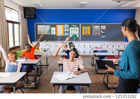 In school, students raising hands in classroom while Indian female teacher with tablet instructing 125353282
