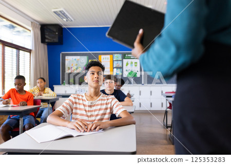 In school, students sitting at desks listening to Indian female teacher holding tablet 125353283