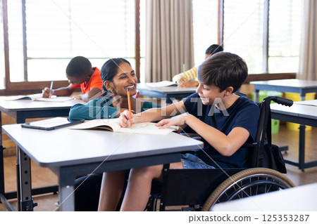 In school, Indian female teacher assisting student in wheelchair with writing in classroom 125353287