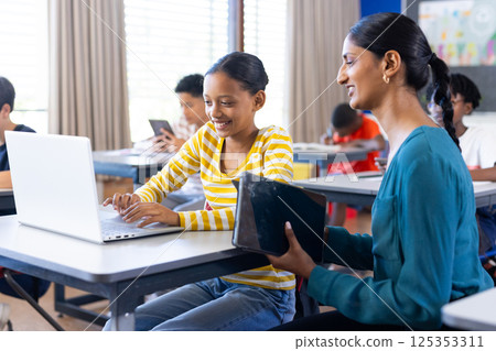 In school, Indian female teacher helping student using laptop in classroom, smiling together In school, Indian female teacher helping student using laptop in classroom, smiling together 125353311