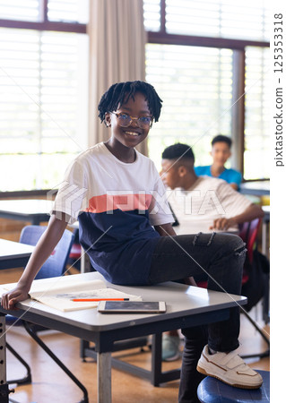In school, smiling student sitting on desk with tablet and notebook nearby In school, smiling student sitting on desk with tablet and notebook nearby 125353318