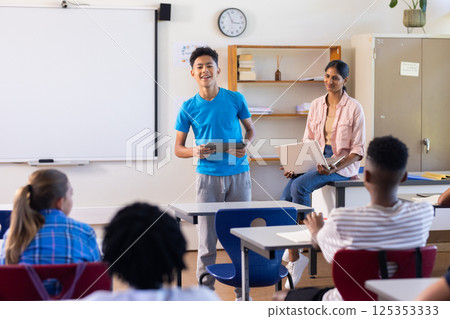 Presenting project in school, boy holding tablet while Indian female teacher and students watching 125353333