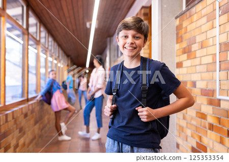 Smiling boy with backpack standing in school hallway with classmates in background 125353354
