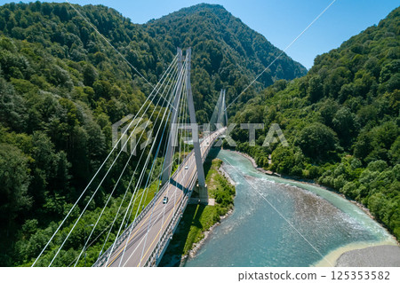 A suspension bridge stretches across a flowing river, surrounded by lush green mountains A suspension bridge stretches across a flowing river, surrounded by lush green mountains 125353582