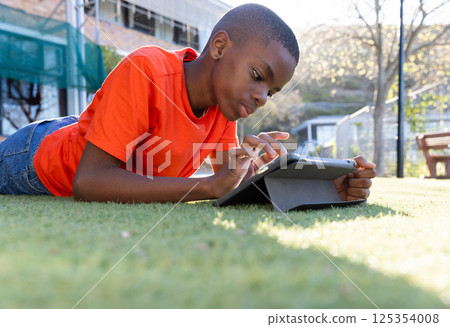 Using tablet, boy lying on grass in school, focusing on screen 125354008