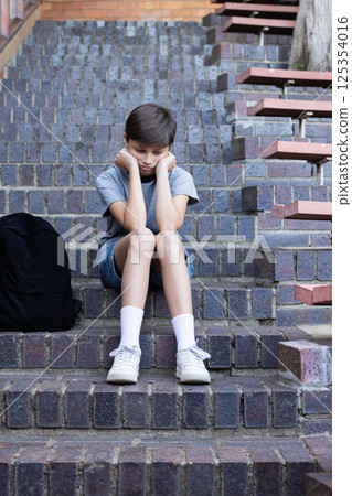 Sitting on school steps, boy looking down with backpack beside him Sitting on school steps, boy looking down with backpack beside him 125354016