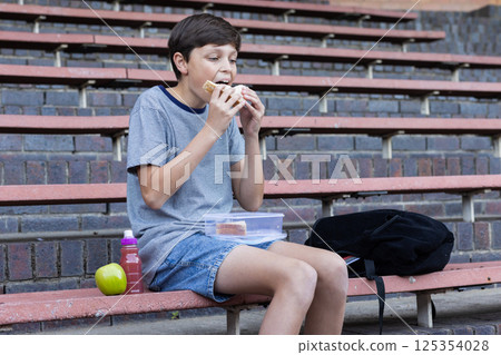 Eating sandwich, boy sitting on bleachers with lunchbox, apple, and backpack 125354028