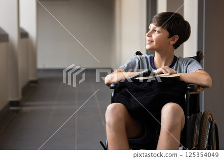 In school, boy in wheelchair holding book, smiling and looking away, copy space 125354049