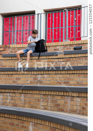 Sitting on steps, boy opening backpack in school with red lockers behind, copy space Sitting on steps, boy opening backpack in school with red lockers behind, copy space 125354057