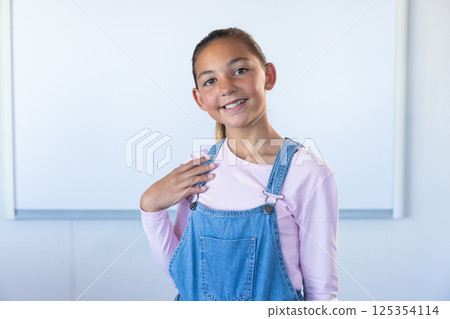 Smiling girl in school classroom wearing denim overalls and pink shirt 125354114