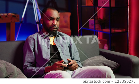 Focused gamer in RGB lit home gaming with gamepad, enjoying himself. Man sitting on sofa in neon illuminated living room, playing videogames on console using controller, camera A 125356273