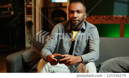 Man arriving home, gaming and enjoying leisure time. Man sitting on couch in neon illuminated living room, playing videogame on console, relaxing after hard day at work, camera A Man arriving home, gaming and enjoying leisure time. Man sitting on couch in neon illuminated living room, playing videogame on console, relaxing after hard day at work, camera A 125356274