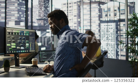 Employee with grimace on face stretches to relieve muscle strain while conducting code review to fix bugs. Fatigued staff member in office suffers back pain from bad posture, optimizing code, camera B 125356322