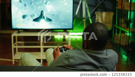 Man playing SF videogames on gigantic smart TV, relaxing after long day at work. Competitive gamer having fun on gaming console in apartment, piloting spaceship using controller, camera B 125356333