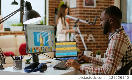 African american photo editor retouches images on his computer in a tech inspired workspace. Man does freelance tasks for photo retouching and digital art, media and graphic design sectors. Camera B. African american photo editor retouches images on his computer in a tech inspired workspace. Man does freelance tasks for photo retouching and digital art, media and graphic design sectors. Camera B. 125356340