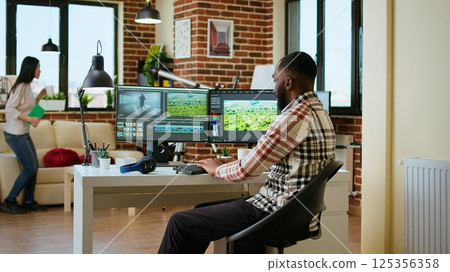 Male video editor works from modern home office in the living room, multitasking with a dual monitor setup and editing software. Finishing post production tasks for media production. Camera A. 125356358