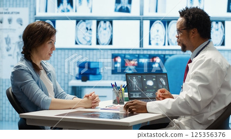 Expert evaluating diagnostic results with a patient in a medical cabinet, showcasing advanced healthcare technology using a screen displaying x rays and MRI scans. Care management. Camera A. 125356376