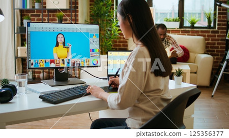 Asian graphic designer uses editing software for photo retouching in a well organized home workspace. Female freelancer uses digital tools essential for a creative artistic career. Camera A. 125356377