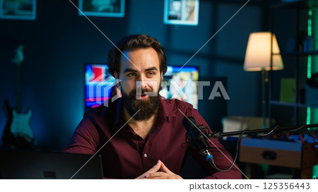 Joyful content creator sitting at table talking in microphone in dimly lit home studio interior, chatting with subscribers. Upbeat man surrounded by podcast recording gear in apartment, camera A 125356443
