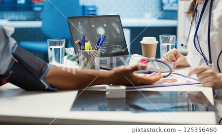 Female medic measuring blood pressure and pulse for a patient, working with blood pressure monitor and other diagnostic tools. Professional cardiology examination, high quality readings. Camera A. 125356460