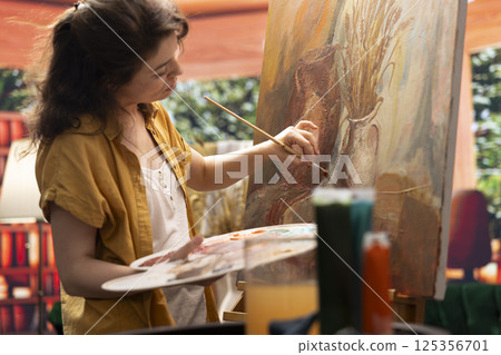 Female painter using watercolor paint and mixing colors on palette for her artistic creation in fine art workshop. Woman painting rustic elements on canvas, creative process in atelier. 125356701